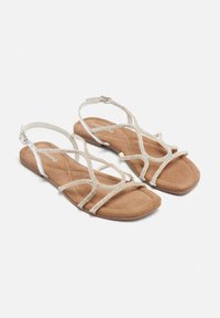 Sandali piatti con una soletta in ecopelle scamosciata color tan, cinturini decorativi argentati con accenti glitterati e una fibbia alla caviglia regolabile.