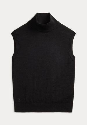 Femme debout les mains dans les poches, vêtue d’un col roulé noir sans manches, d’un pantalon large gris avec une ceinture noire, de chaussures noires et d’un sac à bandoulière.