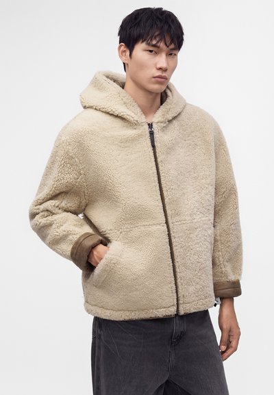 Man wearing beige fuzzy hooded jacket with front zipper and side pockets, paired with black pants, standing against plain white background.