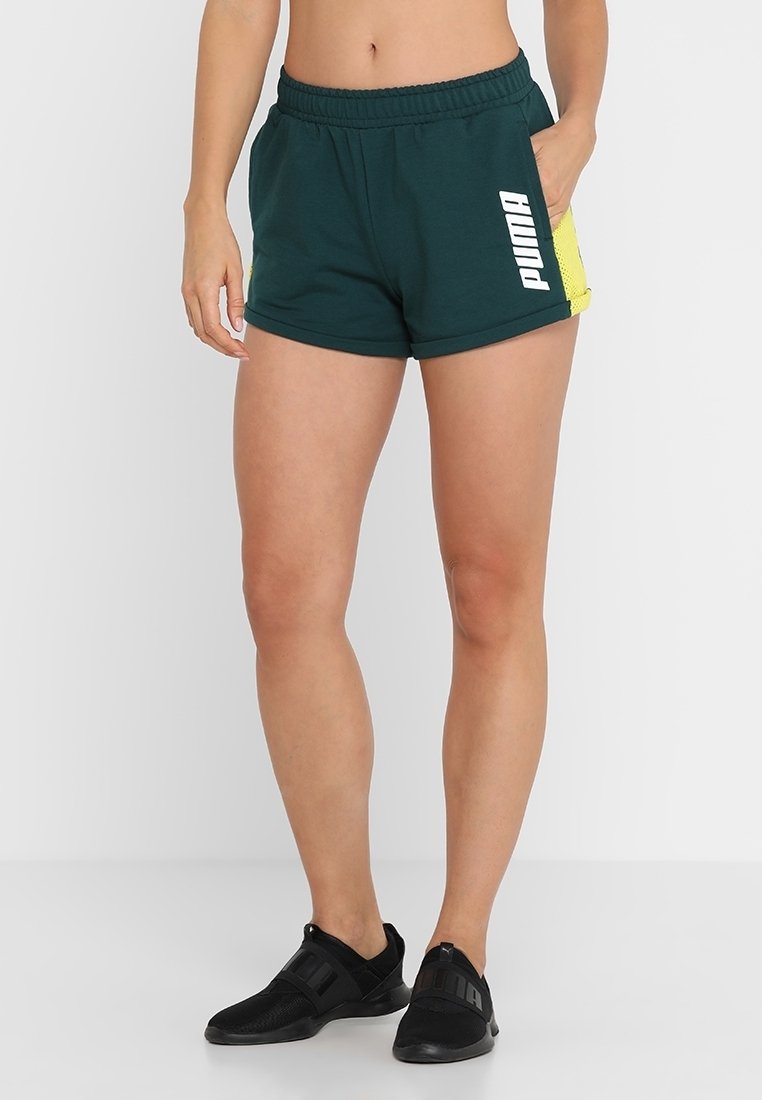Mujer vestida con pantalones cortos Puma de color verde oscuro con acentos amarillos en los laterales y zapatillas deportivas negras de tipo slip-on, de pie frente a un fondo blanco liso.