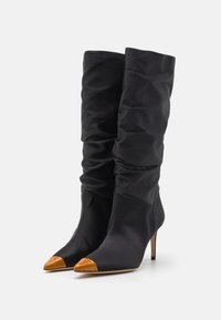 Black satin knee-high boots with a slouchy design, pointed toe, and glossy tan cap toe. Stiletto heel adds elevation.