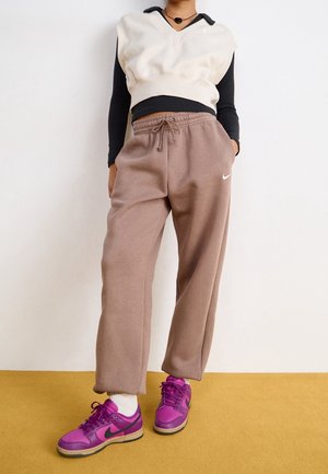 Person, die braune Nike-Sweatpants mit Kordelzug, weiße Socken und violette Nike-Sneaker trägt, steht auf gelbem Boden vor einer weißen Wand.