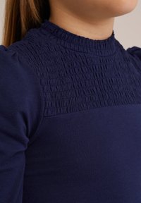Tamsiai mėlyna bliuska su raukšlėtu kaklu ir pūkuotomis rankovėmis. Audinys turi minkštą tekstūrą, prigludusią siluetą ir apvalų iškirpimą.