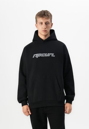 Young man wearing a black hoodie with white "rip curl" logo, standing against a plain white background.