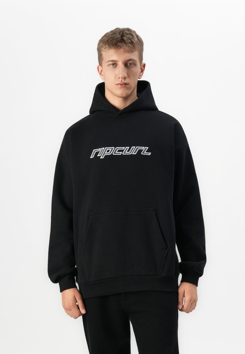 Mladý muž v čiernej mikine s kapucňou s bielym logom "rip curl", stojaci na jednofarebnom bielom pozadí.