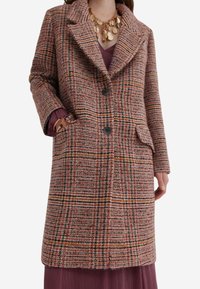 Kvinde iført en midtlængde pink-orange ternet frakke med sorte knapper, en lilla kjole og en chunky guldhalskæde, hånd i lommen.