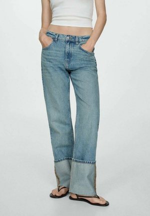 Woman wearing light blue straight-leg jeans with folded cuffs, white cropped tank top, and black flip-flops, hands in pockets, standing on plain background.