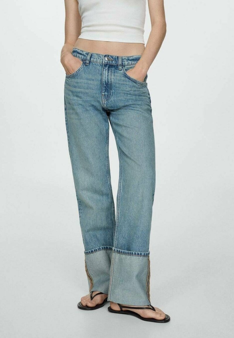 Femme portant un jean droit bleu clair avec revers pliés, un débardeur blanc court et des tongs noires, les mains dans les poches, debout sur un fond uni.