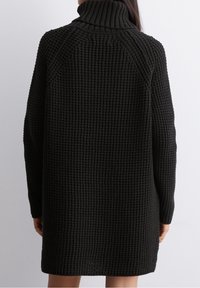 Robe en tricot noir avec un col montant, un motif texturé et des manches longues. Présente une coupe ample et des détails côtelés au niveau du cou.