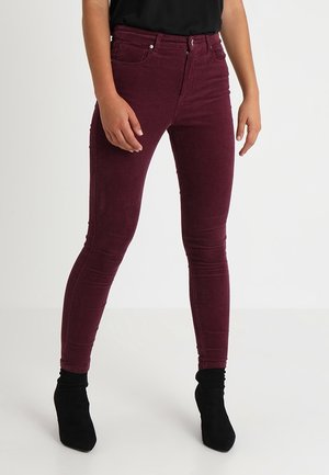 Frau trägt schlanke, hoch taillierte burgunderrote Cordhosen und schwarze Knöchelstiefel, steht vor einem einfachen weißen Hintergrund.