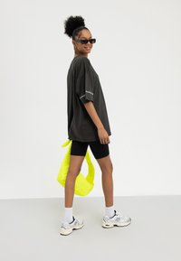 Frau trägt übergroßes schwarzes T-Shirt und Radlerhosen, weiße Turnschuhe, schwarze Sonnenbrille, hält neon-gelbe gesteppte Tasche, lächelt vor weißem Hintergrund.