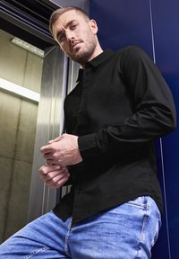 Zwarte blouse met lange mouwen en kraag, gecombineerd met lichtblauwe jeans. Model poseert tegen een metalen liftachtergrond.