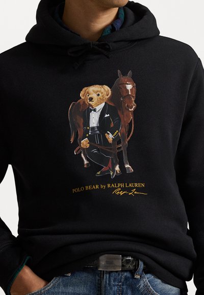 Juodas bliuzonas su grafika, kurioje pavaizduotas lokys smokinge šalia rudo arklo. Dizainą puošia logotipas ir parašas geltonomis raidėmis.