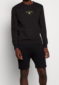 Svart bomullströja med långa ärmar, rund hals och gul "NEW HORIZON"-text. Matchas med svarta shorts med en enkel design.