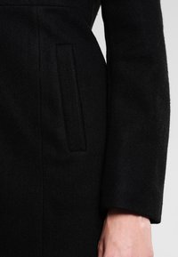 Abrigo de lana negro con un bolsillo lateral y una textura suave. La manga es larga y presenta un acabado limpio y a medida.