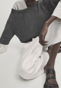 Gebreide oversized grijze trui met een brede neklijn, gelaagd over een gestreept wit shirt en gecombineerd met losse witte broeken en zwarte gevlochten sandalen.