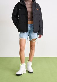 Sort dunjakke med lynlås, blå slidte denimshorts og hvide ankelstøvler med grøn detalje. Strukturerede materialer og unikt design.