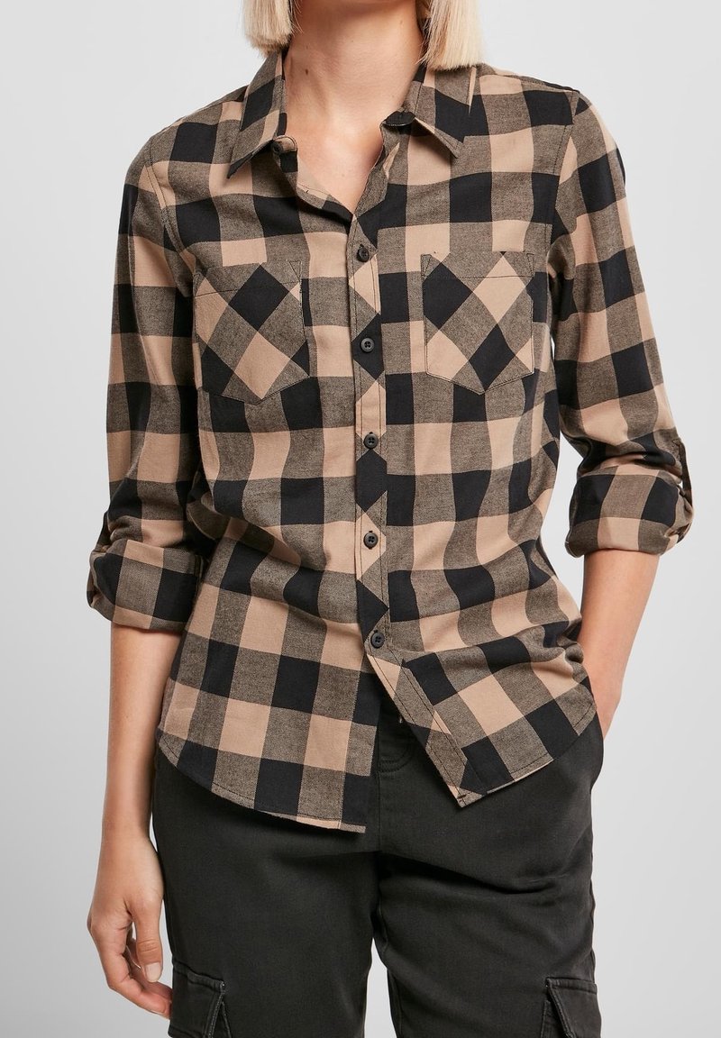 Femme portant une chemise à manches retroussées à carreaux beige et noirs avec deux poches poitrine, associée à un pantalon cargo noir.