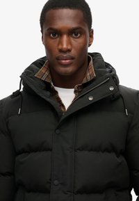 Czarna kurtka puchowa z kapturem i sznurkami, pod którą znajduje się koszula w kratę oraz biała koszulka. Gładka tekstura z zapięciem na guziki.