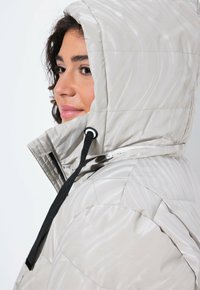 Chaqueta acolchada de color gris claro con capucha, que presenta una textura ondulada, cordones elásticos y un patrón acolchado. Detalle de cremallera en el cuello.