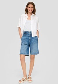 Weißes kurzärmliges Hemd mit Lochmuster, kombiniert mit hellblauen Denim-Shorts. Das Model trägt beige Sandalen und steht vor einem grauen Hintergrund.