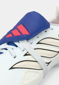 Sportssko med hvidt syntetisk overmateriale, blå polstret tunge, røde accenter og sorte kurvede mønstre. Har snørebånd og en tekstureret overflade.