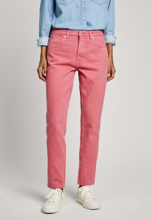 Person, die pinke Jeans mit geradem Bein, hellblaues hochgekrempeltes Langarmhemd und weiße Schnürsneaker trägt, steht vor einem einfarbigen Hintergrund.