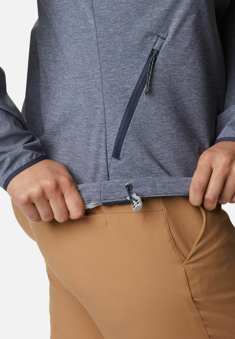 Giacca leggera in tessuto blu con tasca con zip e orlo regolabile. Abbinata a pantaloni attillati color sabbia, che mostrano una texture e una vestibilità lisce.