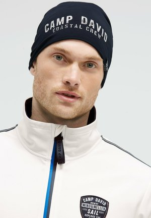 Man wearing navy beanie with "CAMP DAVID COASTAL CREW" text and white zip-up jacket featuring a Camp David logo patch.