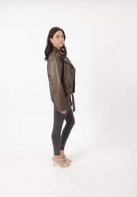 Veste de moto en faux cuir marron avec un design court, de grands revers et une fermeture à pression, associée à un jean skinny gris et des talons légers.