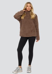 Bruin fleece pullover met een kwart-rits, met een gestructureerde oppervlakte en zijzakken, gecombineerd met zwarte legging en zilveren sportschoenen.