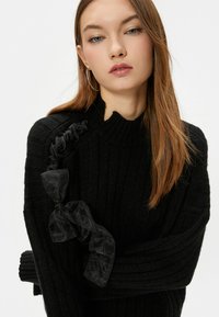 Czarny sweter ribbed z wysokim kołnierzem, z teksturowanym designem oraz czarną przezroczystą kokardą akcentującą na lewym ramieniu.