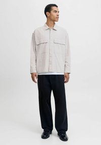 Camicia oversize grigia chiara con motivo a griglia, dotata di due tasche sul petto e chiusura con bottoni, abbinata a pantaloni neri a gamba larga e scarpe nere.