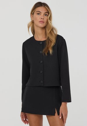 Giacca nera corta con maniche lunghe, scollatura rotonda e sei bottoni decorativi. Abbinata a una gonna nera coordinata con uno spacco laterale.