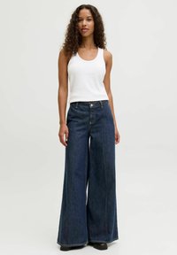 Woman with curly hair wearing a white sleeveless top and wide-leg dark blue jeans, standing against a plain light background.