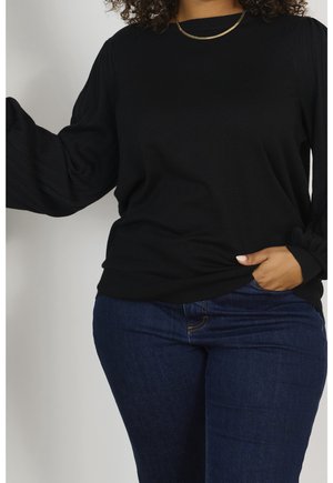 Maglione nero con maniche a coste testurizzate e scollatura rotonda, abbinato a jeans blu scuro. Il materiale sembra morbido e confortevole. Collana dorata visibile.