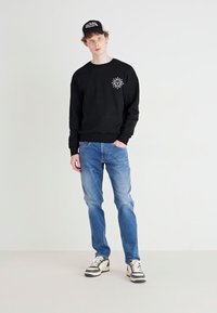Black sweatshirt with a circular sun graphic; blue denim jeans; black and white sneakers with striped accents; baseball cap.