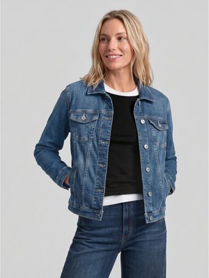 Une femme souriante pose, les mains dans les poches. Elle porte une veste en jean bleue et un jean par-dessus un haut noir, sur fond de studio gris.