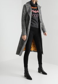 Šedý kostkovaný kabát s klopami, černý rolák se barevnými vzory, černé úzké džíny a černé boty na podpatku.