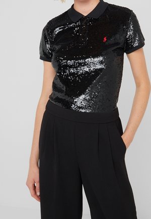 Woman wearing a short-sleeve black sequin polo shirt with a red logo paired with high-waisted black pants, hand in pocket.