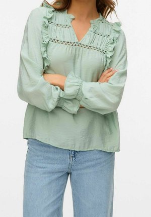 Woman wearing a light green blouse with ruffles and lace details, paired with light blue jeans, arms crossed over chest.