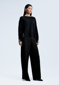 Černý pletený svetr s uvolněným střihem a bočními akcenty. V kombinaci s černými kalhotami s vysokým pasem, které mají strukturovaný povrch a široké nohavice.