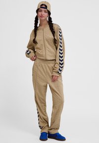 Noor naine palmikutega, kes kannab beežist dressi tumeliste siniste chevrontriipudega, beeži mütsi ja siniste tossudega, seisab valgel taustal.