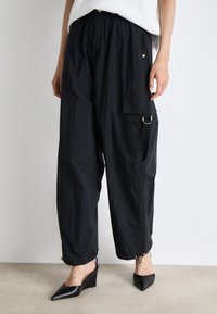 Pantalons cargo légers et noirs avec une taille élastique, des poches latérales et des cordons de serrage aux chevilles. Portés avec des chaussures à talons compensés noires.