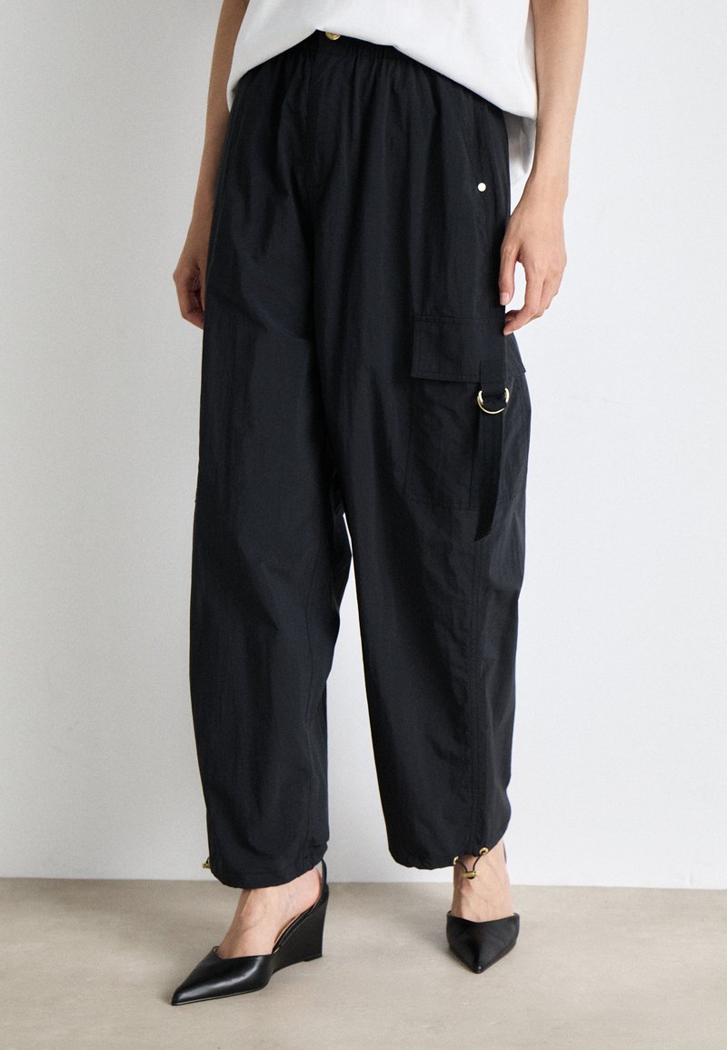Pantalons cargo légers et noirs avec une taille élastique, des poches latérales et des cordons de serrage aux chevilles. Portés avec des chaussures à talons compensés noires.