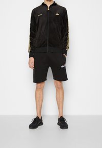 Svart sammet zip-up jacka med guldaccessoarer, kombinerad med svarta shorts med en logotyp. Modellen bär svarta träningsskor.