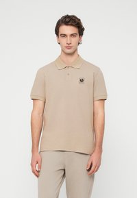 Polo beige avec une texture subtile, doté d'un col et d'une patte de boutonnage à trois boutons. Un petit écusson logo noir est placé sur la poitrine.