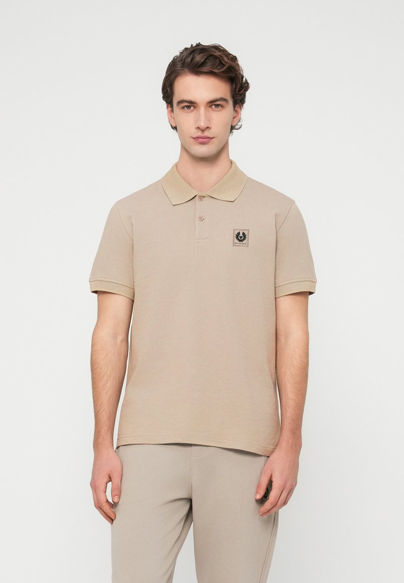 Polo beige avec une texture subtile, doté d'un col et d'une patte de boutonnage à trois boutons. Un petit écusson logo noir est placé sur la poitrine.