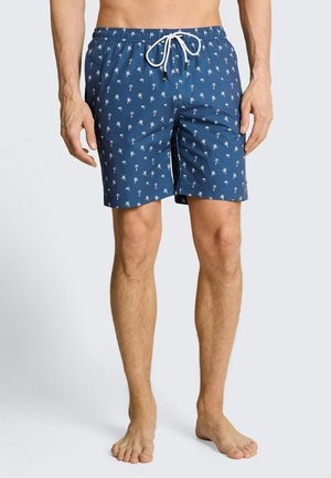 Hombre con pantalones de baño azules con estampado blanco de palmeras y cordón blanco, de pie descalzo sobre un fondo neutro.