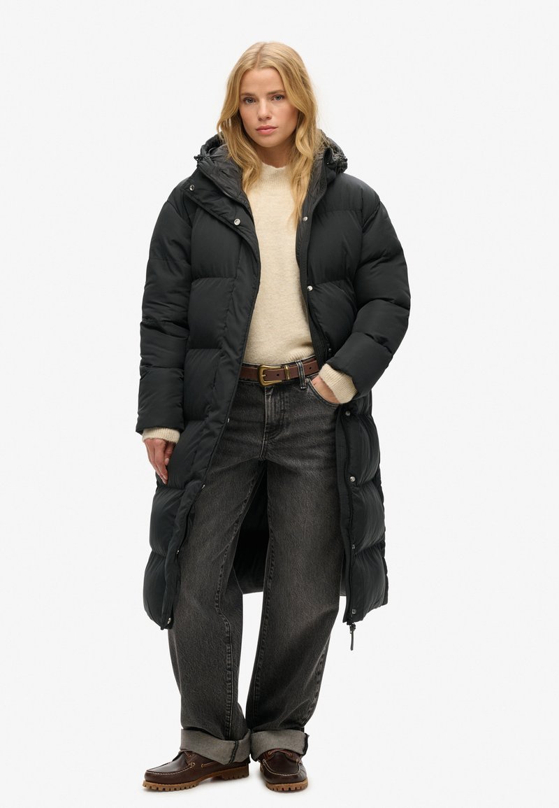 Sort pufferjakke med hætte, med en quiltet design, sidelommer og lang længde. Modellen har en beige sweater og mørke jeans på.
